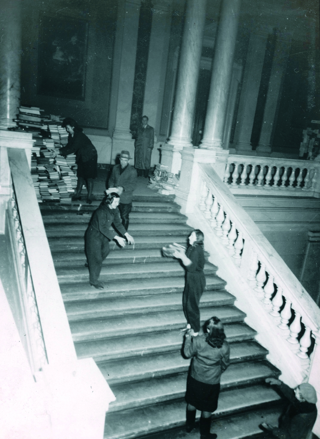 Fotografie der Bestandsauslagerung von Bibliotheksbüchern: Personen reichen sich Buchbestände auf den Treppenstufen der Albertina untereinander weiter, im Hintergrund sind große Stapel Bücher zu sehen