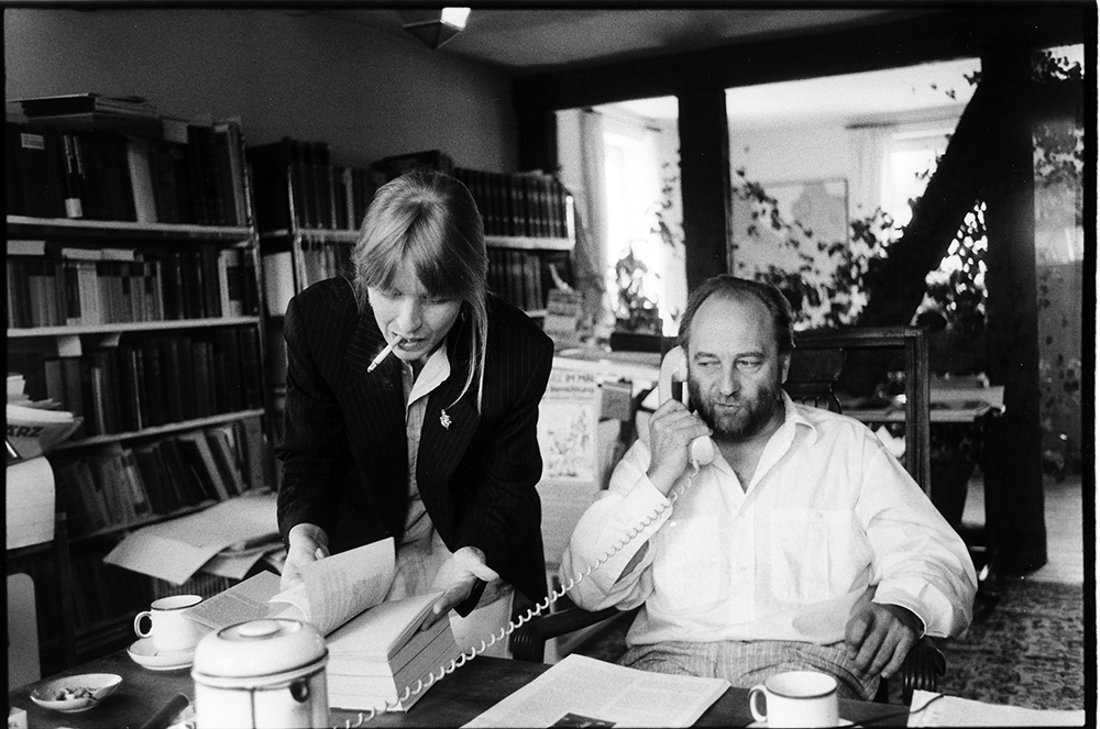 Barbara Kalender und Jörg Schröder während der Arbeit an einem Schreibtisch. Barbara Kalender raucht und blättert in einem Buch. Jörg Schröder telefoniert.